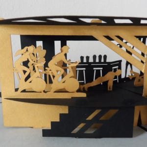 Peut inclure: Carte pop-up repr&eacute;sentant une salle de sport, fabriqu&eacute;e en papier noir et dor&eacute;. Des silhouettes de personnes font de l'exercice, notamment du v&eacute;lo, des pompes et de l'halt&eacute;rophilie. La carte a un design en couches, cr&eacute;ant un effet tridimensionnel.