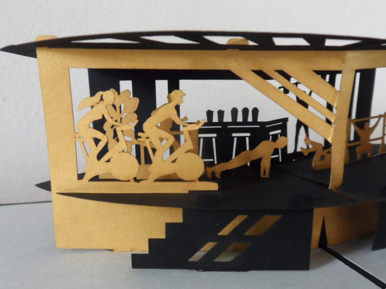 Peut inclure: Carte pop-up repr&eacute;sentant une salle de sport, fabriqu&eacute;e en papier noir et dor&eacute;. Des silhouettes de personnes font de l'exercice, notamment du v&eacute;lo, des pompes et de l'halt&eacute;rophilie. La carte a un design en couches, cr&eacute;ant un effet tridimensionnel.
