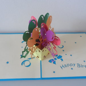 Puede incluir: Una tarjeta pop-up 3D con una caja amarilla llena de globos de colores. La tarjeta est&aacute; abierta y el interior dice "Feliz cumplea&ntilde;os".