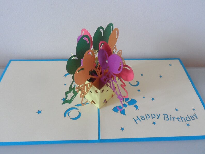 Puede incluir: Una tarjeta pop-up 3D con una caja amarilla llena de globos de colores. La tarjeta est&aacute; abierta y el interior dice "Feliz cumplea&ntilde;os".
