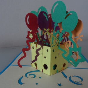 Puede incluir: Una caja amarilla con un dise&ntilde;o pop-up que presenta globos coloridos, serpentinas y estrellas. La caja est&aacute; abierta y los globos salen de la parte superior. La caja est&aacute; decorada con lunares y un patr&oacute;n en zigzag.