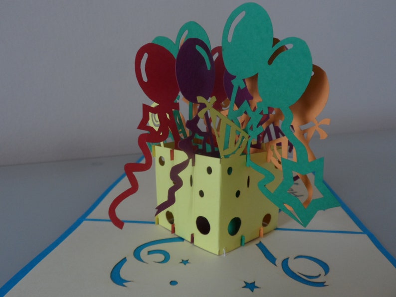 Puede incluir: Una caja amarilla con un dise&ntilde;o pop-up que presenta globos coloridos, serpentinas y estrellas. La caja est&aacute; abierta y los globos salen de la parte superior. La caja est&aacute; decorada con lunares y un patr&oacute;n en zigzag.