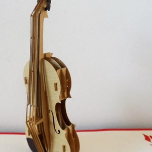 May include: A wooden pop-up card featuring a detailed model of a violin. The violin is made of light brown wood and has a dark brown finish on the bottom.
