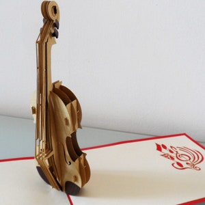 May include: A brown and beige paper pop-up card featuring a 3D violin. The card is open and the violin is standing upright. The background of the card is white with a red border. The inside of the card features a red paper cut-out design of a treble clef.