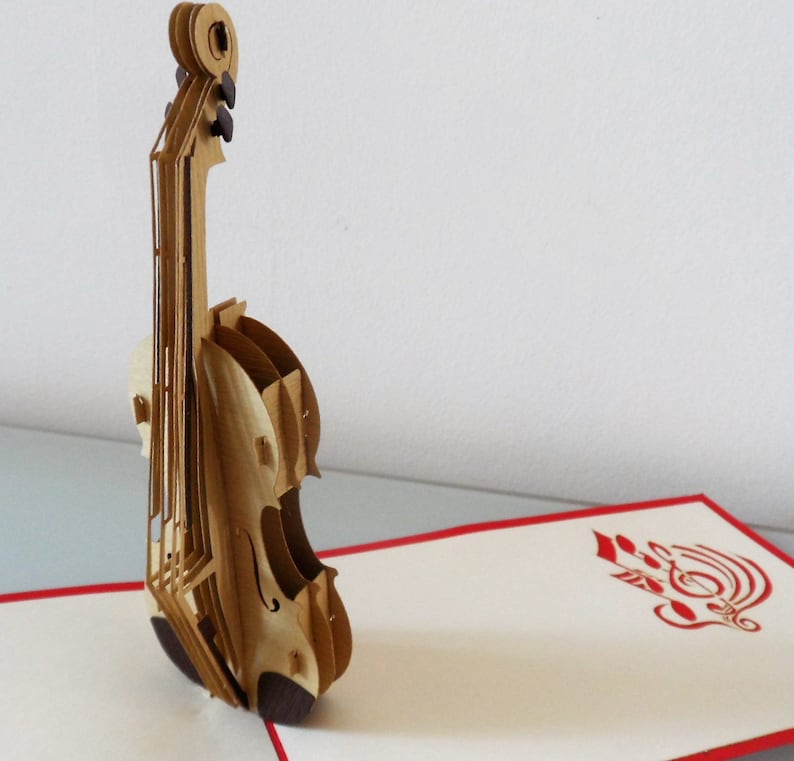 May include: A brown and beige paper pop-up card featuring a 3D violin. The card is open and the violin is standing upright. The background of the card is white with a red border. The inside of the card features a red paper cut-out design of a treble clef.