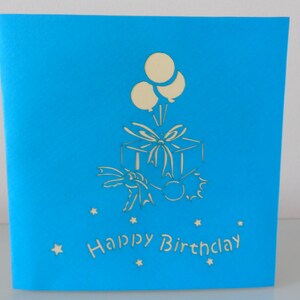 Puede incluir: Una tarjeta azul con un dise&ntilde;o 3D pop-up blanco de una caja de regalo con globos y el texto "Happy Birthday".