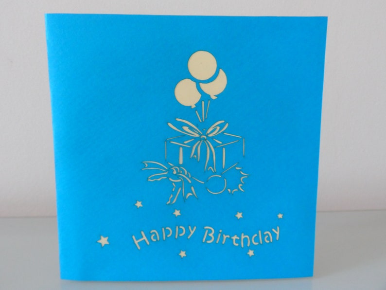 Puede incluir: Una tarjeta azul con un dise&ntilde;o 3D pop-up blanco de una caja de regalo con globos y el texto "Happy Birthday".