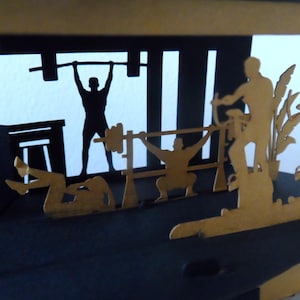 Peut inclure: Une &oelig;uvre d'art en papier en couches repr&eacute;sentant une sc&egrave;ne de salle de sport. Des silhouettes soul&egrave;vent des poids, utilisent des appareils d'exercice et s'&eacute;tirent. L'&oelig;uvre est en noir et or, avec l'accent mis sur les personnages et l'&eacute;quipement.