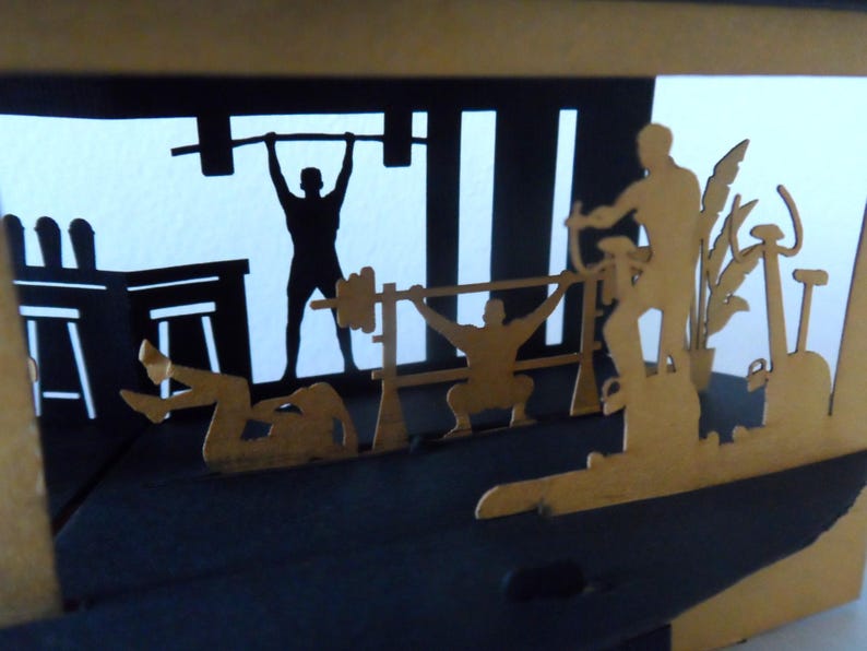 Peut inclure: Une &oelig;uvre d'art en papier en couches repr&eacute;sentant une sc&egrave;ne de salle de sport. Des silhouettes soul&egrave;vent des poids, utilisent des appareils d'exercice et s'&eacute;tirent. L'&oelig;uvre est en noir et or, avec l'accent mis sur les personnages et l'&eacute;quipement.