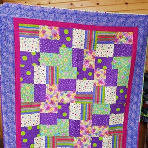 Può includere: Una trapunta patchwork di colore viola, verde, rosa e bianco con un bordo a motivo a spirale. La trapunta è realizzata con diversi tessuti con motivi diversi, tra cui pois, strisce e motivi floreali.