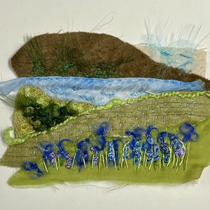 May include: A textile art piece depicting a landscape with a brown hill, a blue river, and a field of blue flowers. The piece is made with different fabrics and textures, creating a layered effect.