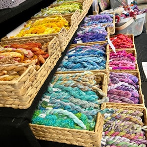 May include: A selection of hand-dyed embroidery floss in various colours is presented in wicker baskets. The floss is arranged in rows, with each row showcasing a different colour gradient. The floss is labelled "Hand dyed floss 12 inch".