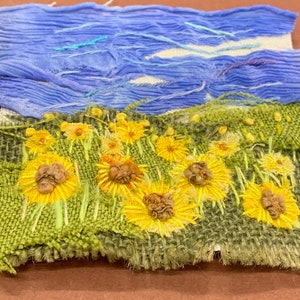 May include: A close-up of a small embroidered artwork depicting a field of yellow flowers against a blue sky. The flowers are stitched with yellow and brown thread, and the sky is made of blue fabric.