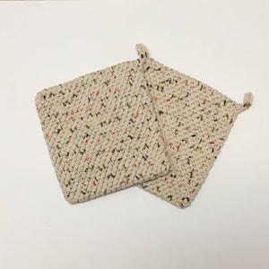 May include: Two beige, square potholders with a speckled pattern of red and green. Each potholder has a small loop in one corner for hanging. The potholders are made of a thick, textured material.