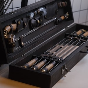 Puede incluir: Una caja de madera con tapa abatible, que contiene un juego de pinchos metálicos y un cuchillo envuelto en cuero. La caja es de madera oscura y tiene un asa en el lateral.