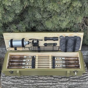 May include: An open, olive-green wooden box with a light wood interior, containing grilling tools. Includes skewers, a metal water bottle, a small grill, a flashlight, and a grey bag. The box is resting on a tree trunk.