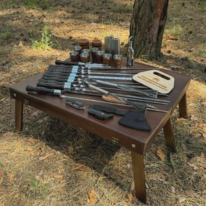 May include: A brown wooden low table displays a collection of outdoor cooking and drinking accessories. Items include skewers, knives, cups, a flask, and a small axe. The items are arranged on a brown table with wooden legs, set outdoors.