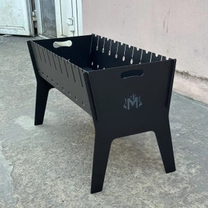 Pode incluir: Churrasqueira de metal preto com um logotipo de uma letra M estilizada com chamas em um suporte com quatro pernas.