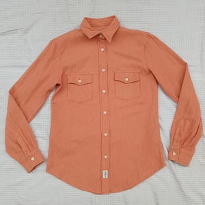 May include: A long-sleeved shirt in a muted orange hue. It has a button-down front, a classic collar, and two chest pockets with buttoned flaps. The cuffs are buttoned. The fabric has a textured appearance.