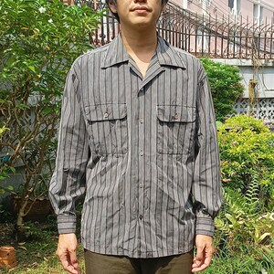 May include: A long-sleeved button-up shirt with vertical stripes in shades of gray, brown, and white. The shirt has two chest pockets and a camp collar. The model is wearing the shirt.