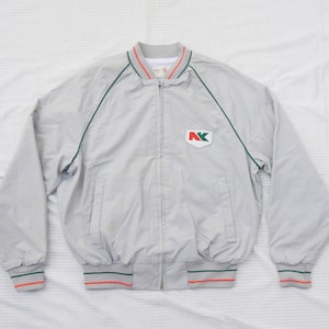May include: A gray bomber jacket with green and red trim. The jacket has a zipper closure and a patch with the letters "NK" in green and red on the left chest.