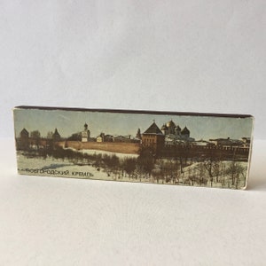 Könnte beinhalten: Eine Vintage-Streichholzschachtel mit einer farbenfrohen Illustration des Nowgorod-Kremls, einer historischen Festung in Russland. Die Streichholzschachtel ist braun mit einem schwarzen Streifen und dem Text "Новгородский Кремль" auf der Vorderseite.
