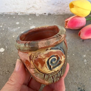 May include: Handmade ceramic pot with a rustic, textured finish. The pot features a spiral design in shades of blue, brown, and yellow. The rim has a green and brown trim. The pot is ideal for small plants or as a decorative item.