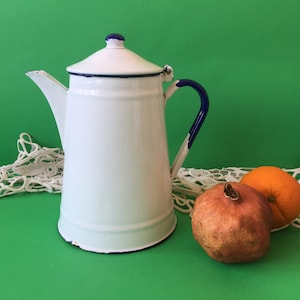 Pode incluir: Bule de esmalte branco com borda e alça azuis. A bule está sobre uma toalha de renda branca com uma romã e uma laranja.