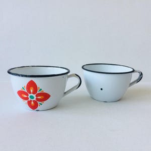 Könnte beinhalten: Zwei weiße Emaille-Tassen mit schwarzen Rändern und Henkeln. Eine Tasse hat ein rotes, orangefarbenes und grünes Blumendesign. Die andere ist weiß mit einem schwarzen Punkt. Vintage-Tassen für Tee oder Kaffee.