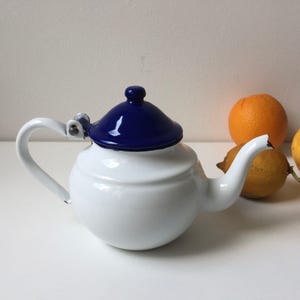 Pode incluir: Uma chaleira de esmalte branco com corpo arredondado e bico curvo. A tampa é azul escuro e a alça é branca. Laranjas e limões ao fundo. Um item de cozinha clássico.