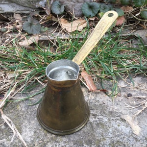 Könnte beinhalten: Eine türkische Kaffeekanne aus Messing mit einem langen Griff. Der Griff ist mit "Elektra, Orvieto" gekennzeichnet.
