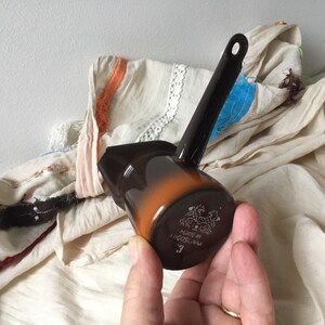 Pode incluir: Uma cafeteira de cerâmica marrom e laranja com uma longa alça preta. A cafeteira tem uma base arredondada e um bico. A parte inferior da cafeteira tem o texto "MADE IN YUGOSLAVIA" e o número "3".
