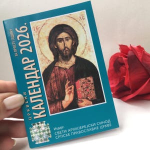Könnte beinhalten: Ein türkisfarbener Kalender für 2026 mit dem Text "ЦРКВЕНИ КАЛЕНДАР 2026." zeigt eine Illustration einer religiösen Figur, die ein Buch hält. Eine rote Rose ist im Hintergrund. Der Kalender wird von einer Person gehalten.