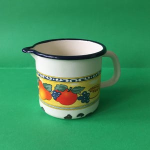 Peut inclure: Pichet en émail blanc avec un bord et une poignée bleus. Le pichet présente une bande jaune avec des illustrations de pommes rouges, de poires et de raisins. Le pichet a un bec verseur.