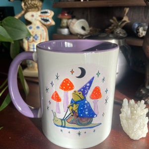 May include: A white ceramic mug with a purple rim and handle. The mug features a whimsical illustration of a frog wearing a wizard hat riding a snail. The frog is holding a wand and the snail is surrounded by mushrooms and stars.