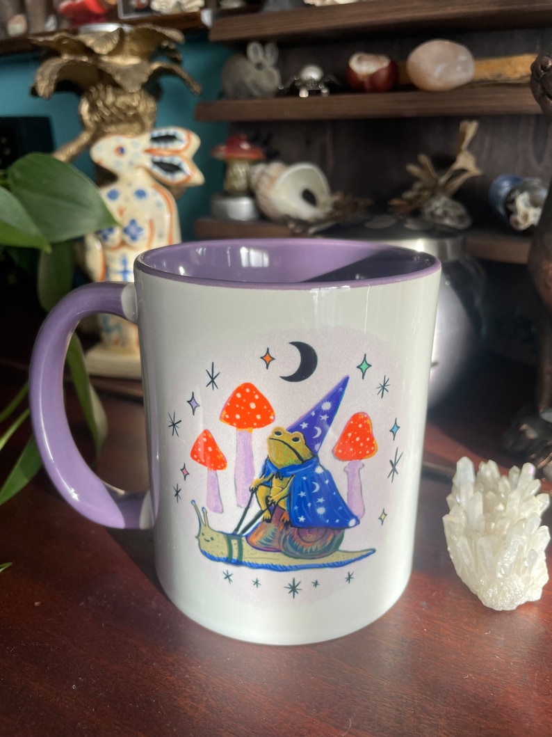 May include: A white ceramic mug with a purple rim and handle. The mug features a whimsical illustration of a frog wearing a wizard hat riding a snail. The frog is holding a wand and the snail is surrounded by mushrooms and stars.