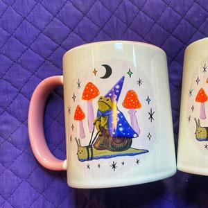 May include: A white ceramic mug with a pink handle features a whimsical illustration of a frog wearing a wizard hat riding a snail. The frog is holding a wand and the snail is surrounded by mushrooms and stars.