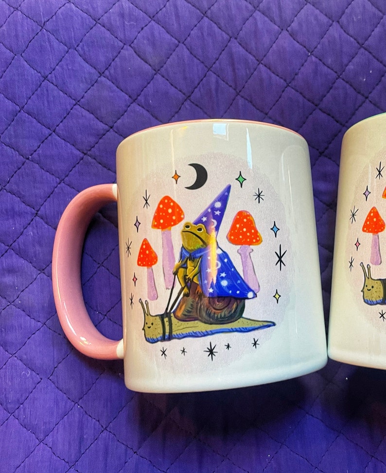 May include: A white ceramic mug with a pink handle features a whimsical illustration of a frog wearing a wizard hat riding a snail. The frog is holding a wand and the snail is surrounded by mushrooms and stars.