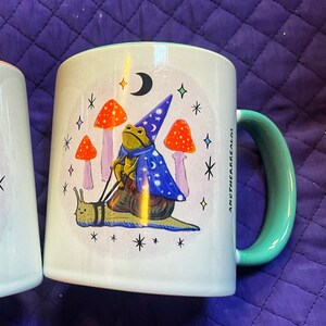 May include: A white ceramic mug with a green rim and handle. The mug features a whimsical illustration of a frog wearing a wizard hat and riding a snail. The frog is holding a wand and there are mushrooms and stars in the background. The text "Another Realm" is printed on the side of the mug.