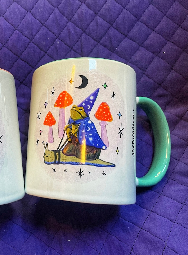 May include: A white ceramic mug with a green rim and handle. The mug features a whimsical illustration of a frog wearing a wizard hat and riding a snail. The frog is holding a wand and there are mushrooms and stars in the background. The text "Another Realm" is printed on the side of the mug.