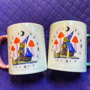 May include: Two white ceramic mugs with pink and green handles. Both mugs feature a whimsical illustration of a frog wearing a wizard hat riding a snail. The frog is holding a wand and there are mushrooms and stars in the background.