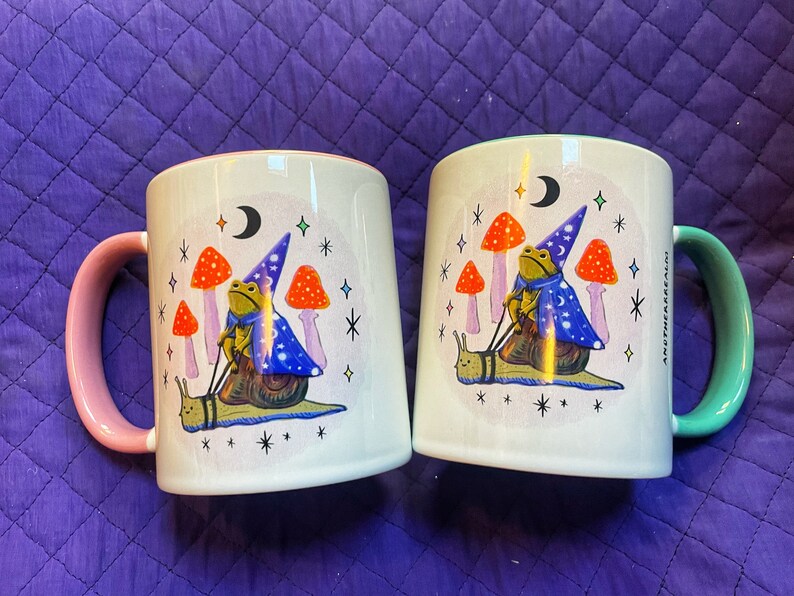 May include: Two white ceramic mugs with pink and green handles. Both mugs feature a whimsical illustration of a frog wearing a wizard hat riding a snail. The frog is holding a wand and there are mushrooms and stars in the background.
