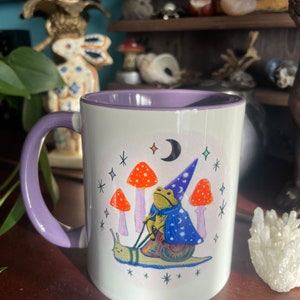 May include: A white ceramic mug with a purple rim and handle. The mug features a whimsical illustration of a frog wearing a wizard hat and riding a snail. The frog is holding a wand and the snail is surrounded by stars and mushrooms.