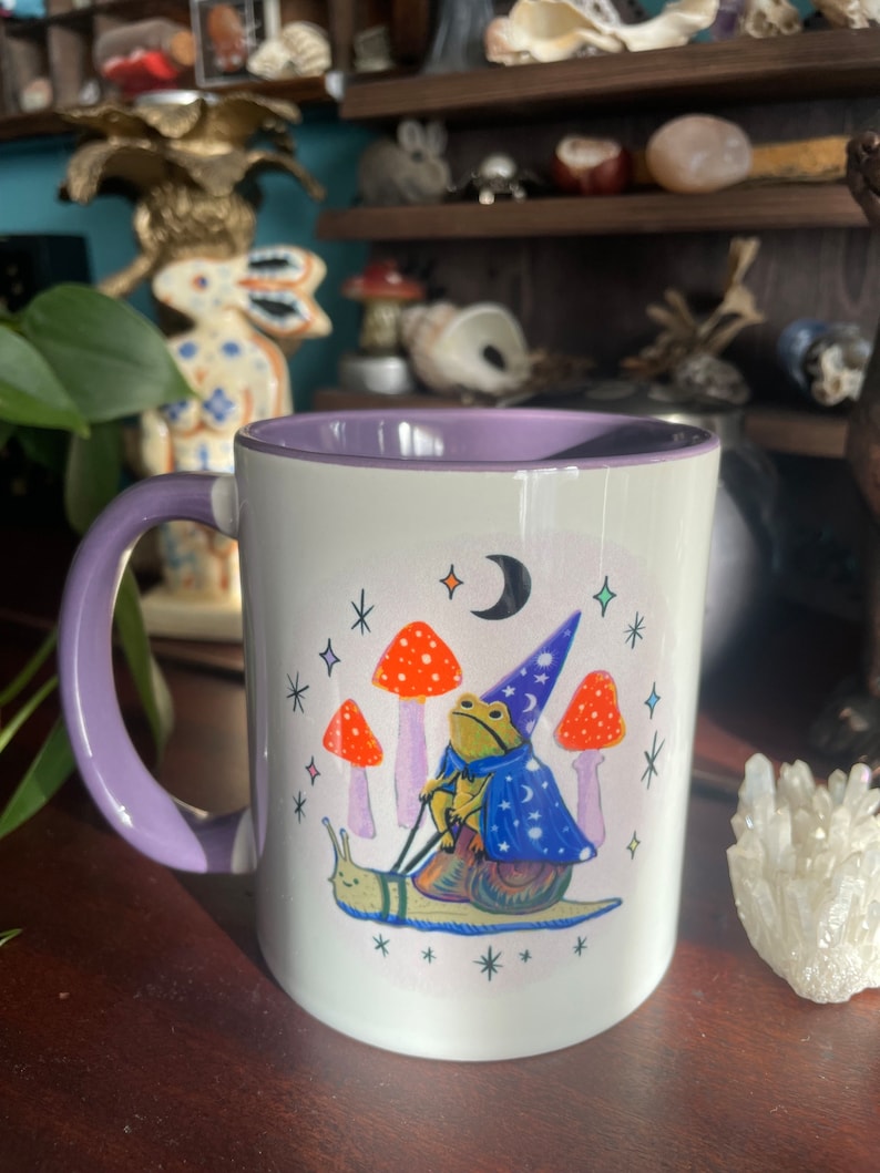 May include: A white ceramic mug with a purple rim and handle. The mug features a whimsical illustration of a frog wearing a wizard hat and riding a snail. The frog is holding a wand and the snail is surrounded by stars and mushrooms.