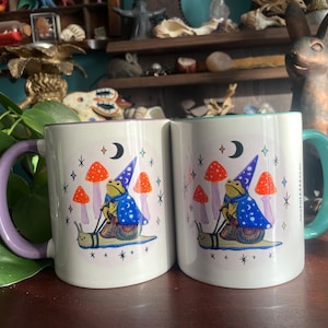 May include: Two white ceramic mugs with a purple and teal rim. Each mug features a whimsical illustration of a frog wearing a wizard hat riding a snail. The frog is surrounded by mushrooms and stars. The text "Magic Happens" is printed on the side of the mug.