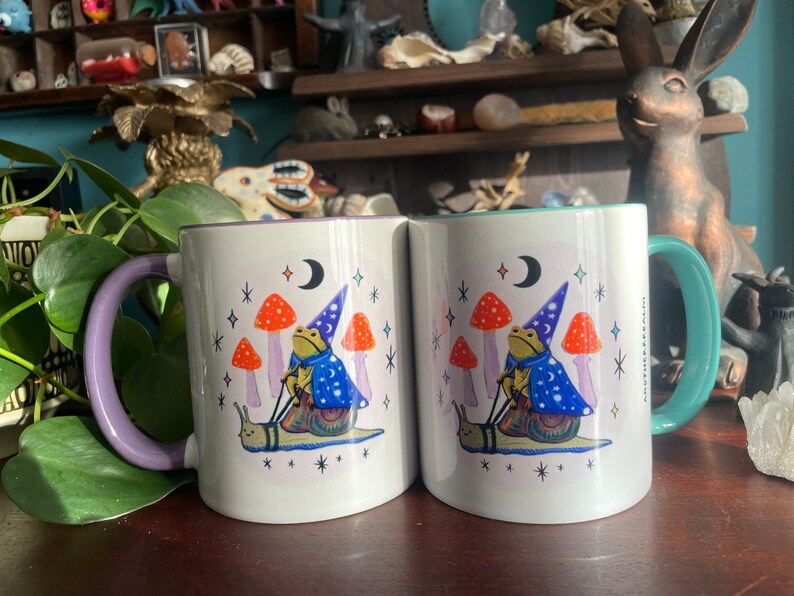 May include: Two white ceramic mugs with a purple and teal rim. Each mug features a whimsical illustration of a frog wearing a wizard hat riding a snail. The frog is surrounded by mushrooms and stars. The text "Magic Happens" is printed on the side of the mug.