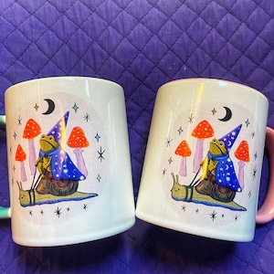 May include: Two white ceramic mugs with a whimsical design of a frog wearing a wizard hat riding a snail. The mugs have a light blue and purple design with a crescent moon and stars. One mug has a teal handle and the other has a pink handle.