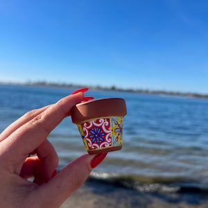 Pode incluir: Um pequeno vaso de flores de terracota com um design decorativo padronizado. O vaso tem uma borda marrom e um padrão vibrante de motivos florais vermelhos, azuis e amarelos. O vaso é segurado contra um fundo de céu azul e água.