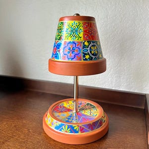 May include: A decorative lamp with a terracotta base and a colorful, patterned shade. The shade features a patchwork design with various floral and geometric motifs in shades of blue, yellow, red, and green. The lamp has a brass stem and a matching base.