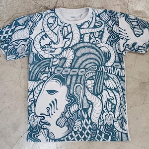 May include: White t-shirt with a teal abstract design featuring a face and intricate patterns. The design covers the entire shirt, including the sleeves. The shirt has a crew neck and short sleeves.